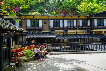 전통적인 일본식 료칸 건물 앞 길가에 앉아 휴식을 취하며 휴대폰을 보고 있는 세 명의 외국인 관광객들과 주변의 푸른 숲 풍경입니다.
