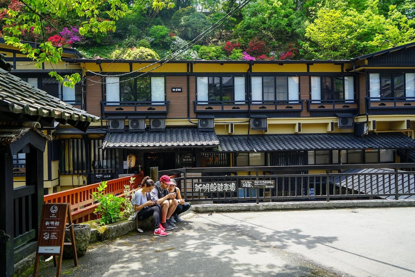 전통적인 일본식 료칸 건물 앞 길가에 앉아 휴식을 취하며 휴대폰을 보고 있는 세 명의 외국인 관광객들과 주변의 푸른 숲 풍경입니다.