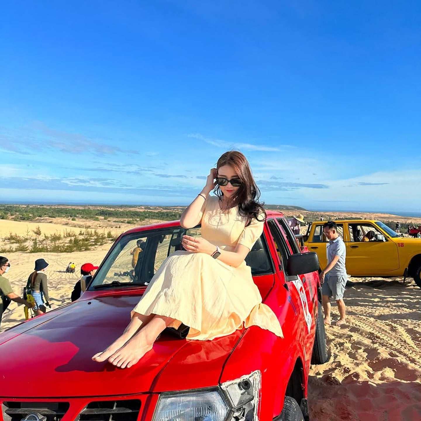 맑고 푸른 하늘이 펼쳐진 사막에서 연한 노란색 원피스를 입은 여성이 빨간색 오프로드 차량 위에 앉아 여유로운 여행의 한때를 즐기고 있습니다.
