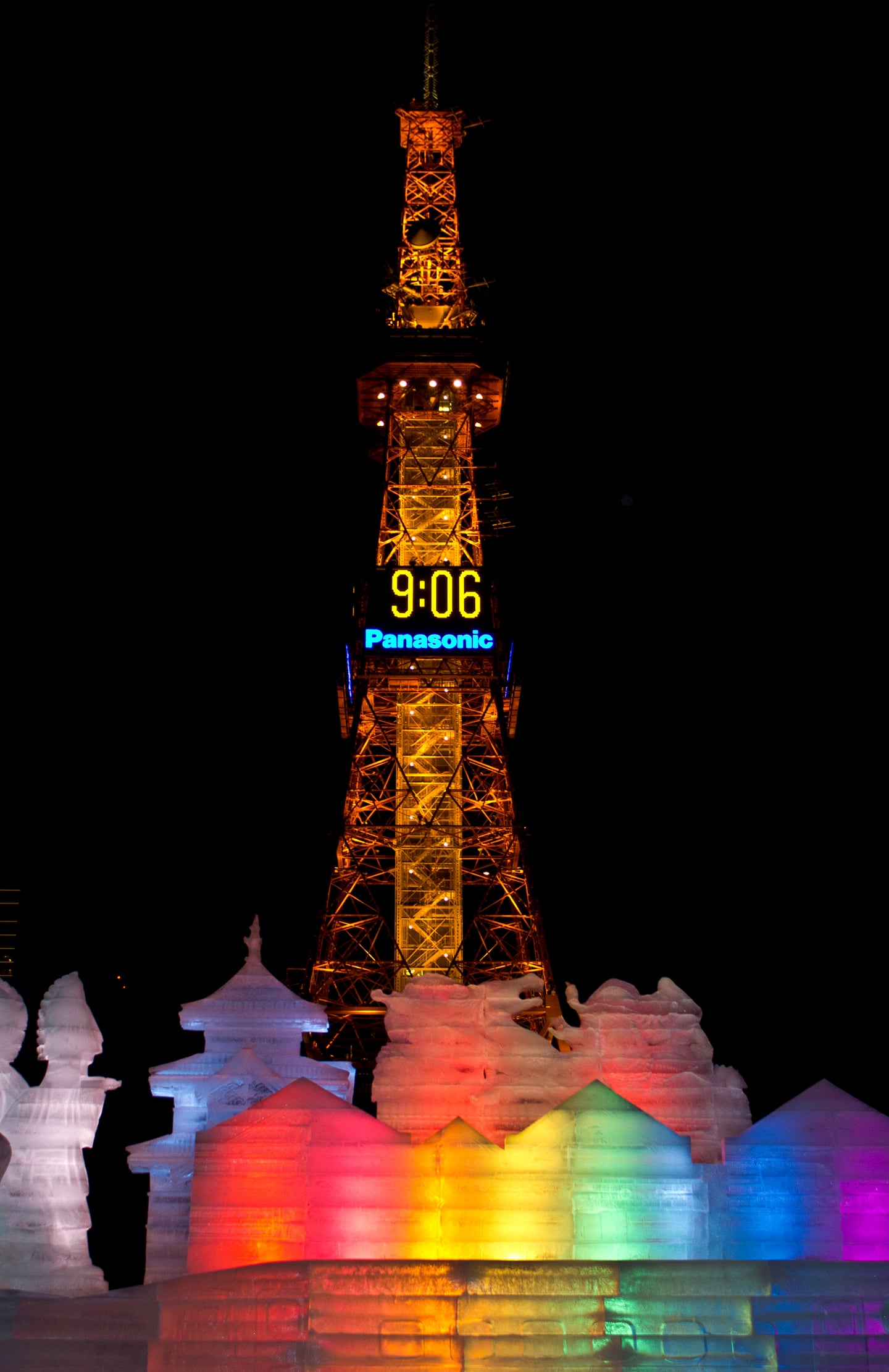 밤하늘 아래 황금빛으로 빛나는 삿포로 TV 타워와 그 앞을 장식한 알록달록한 무지개 빛깔의 얼음 조각들이 어우러진 겨울 축제의 야경입니다.