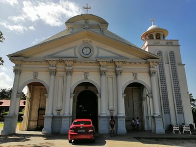 웅장한 기둥과 아치형 입구가 돋보이는 흰색 성당 외관 앞에 빨간색 자동차가 세워져 있고 맑은 하늘이 배경으로 펼쳐진 모습입니다.