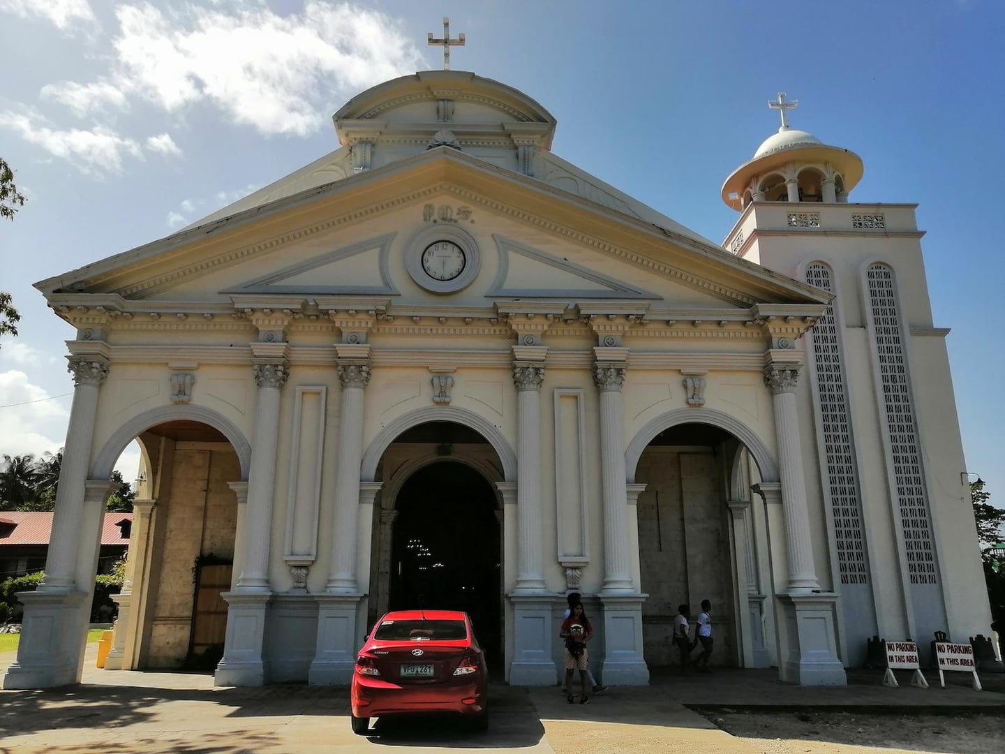 웅장한 기둥과 아치형 입구가 돋보이는 흰색 성당 외관 앞에 빨간색 자동차가 세워져 있고 맑은 하늘이 배경으로 펼쳐진 모습입니다.