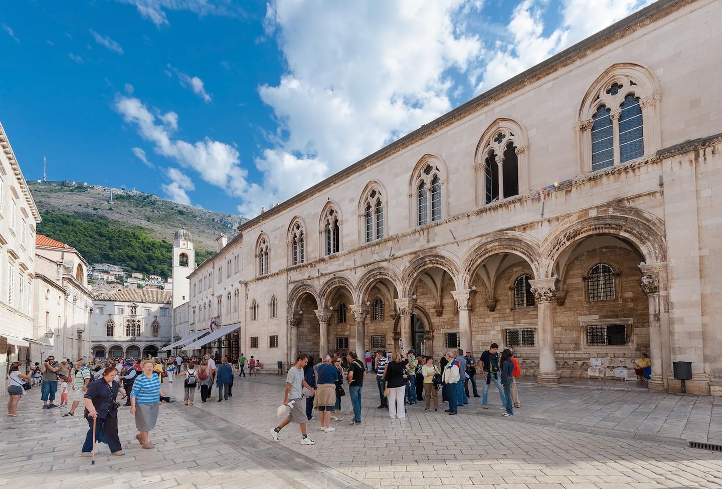 크로아티아 두브로브니크의 렉터 궁전 앞 광장에서 고딕 양식의 아치형 회랑과 석조 건물을 배경으로 산책을 즐기는 관광객들의 활기찬 풍경입니다.