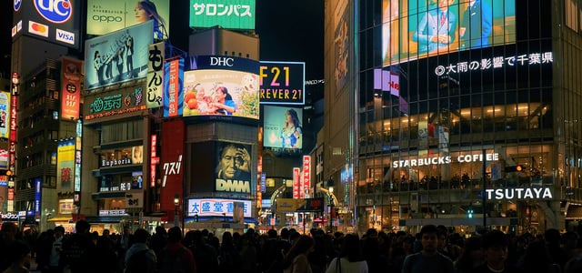 도쿄 시부야의 밤거리를 가득 메운 인파 위로 화려한 대형 전광판과 네온사인 광고들이 빛나며 활기찬 도시의 야경을 보여줍니다.