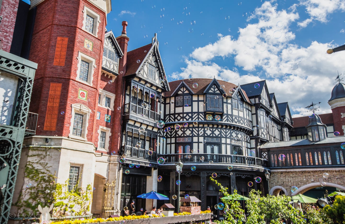 맑은 하늘과 뭉게구름을 배경으로 비눗방울이 흩날리는 이국적인 유럽 스타일의 건축물과 정원이 어우러진 평화로운 야외 전경입니다.