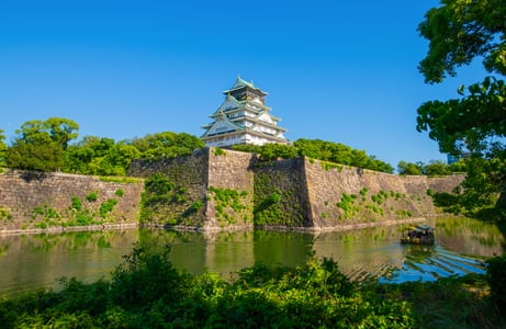 푸른 하늘을 배경으로 견고한 석벽 위에 세워진 오사카 성 천수각과 해자 위를 지나가는 유람선이 어우러진 일본의 역사적인 풍경입니다.