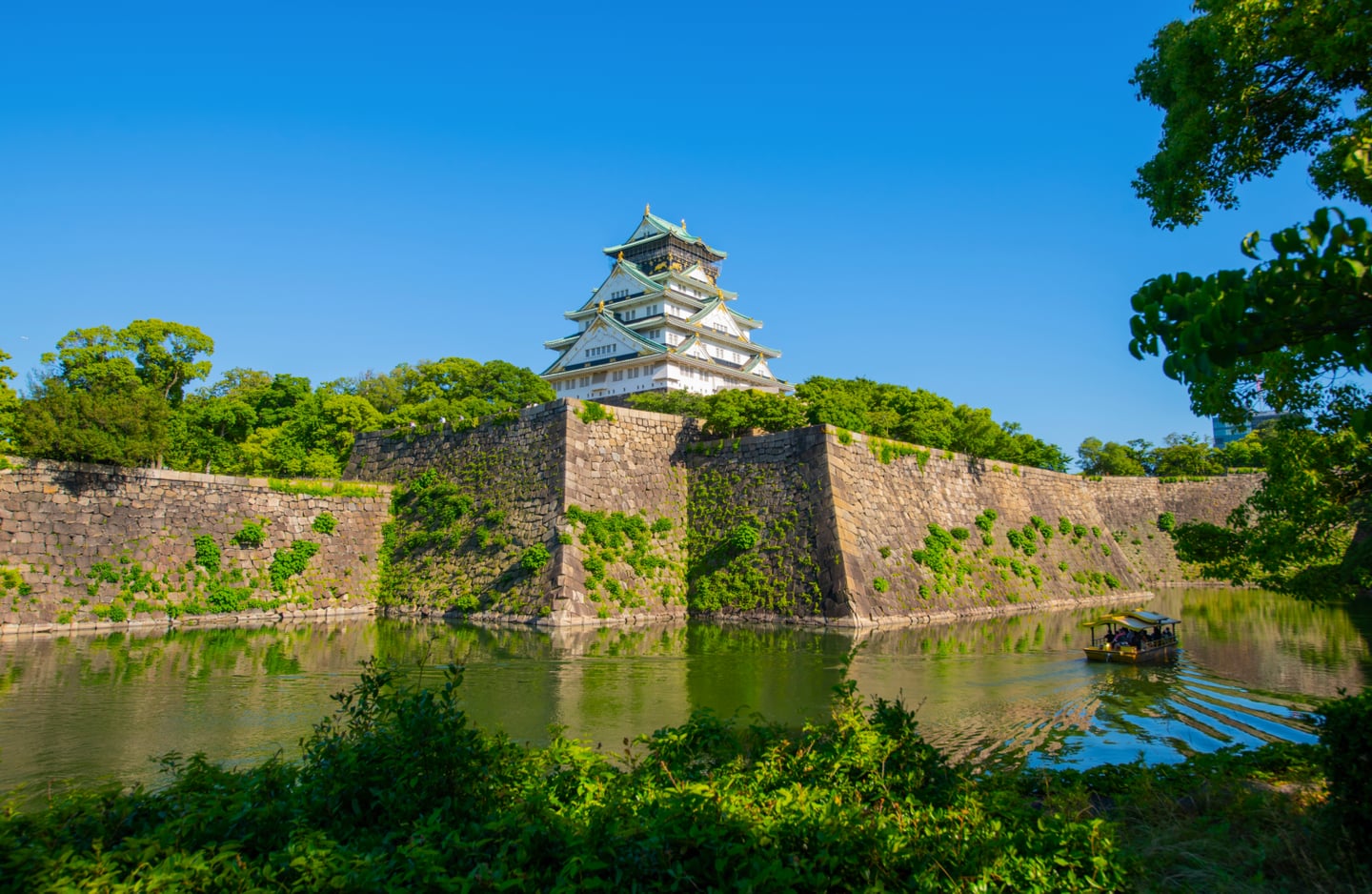 푸른 하늘을 배경으로 견고한 석벽 위에 세워진 오사카 성 천수각과 해자 위를 지나가는 유람선이 어우러진 일본의 역사적인 풍경입니다.