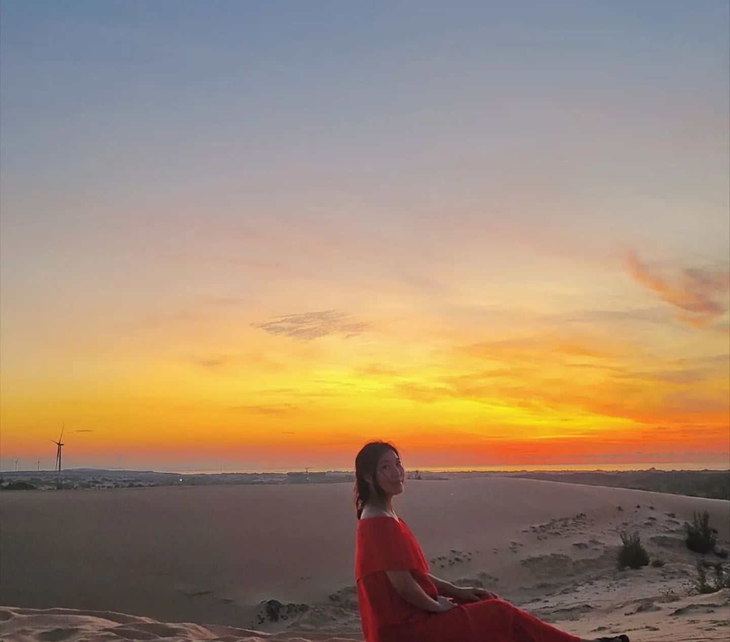 노을이 지는 사막 언덕에 앉아 붉은 드레스를 입고 여유로운 시간을 보내는 여성의 뒷모습과 황금빛 하늘이 어우러진 풍경입니다.