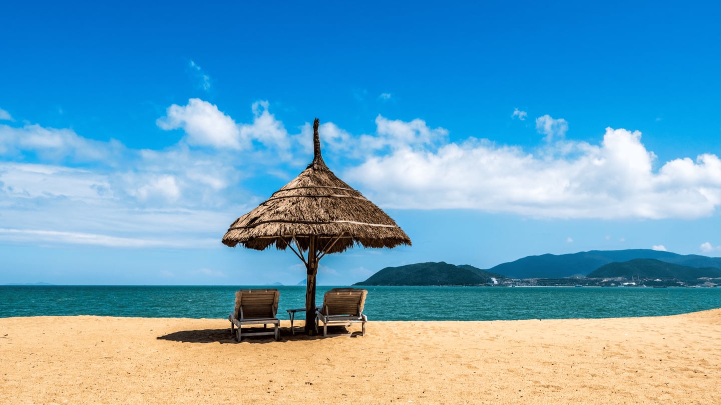 푸른 바다와 하늘을 배경으로 모래사장 위에 세워진 초가 파라솔 아래 나란히 놓인 두 개의 나무 선베드가 평화로운 휴식의 순간을 보여줍니다.