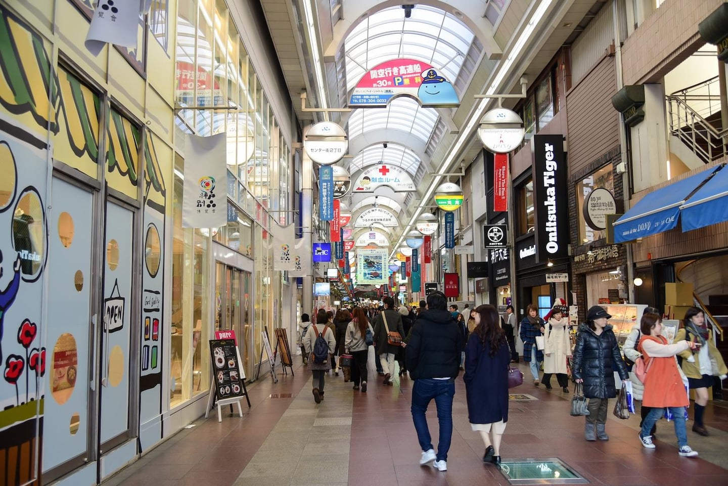 다양한 브랜드 매장과 화려한 간판이 가득한 일본의 지붕 있는 아케이드 쇼핑 거리를 걷고 있는 사람들의 활기찬 풍경입니다.