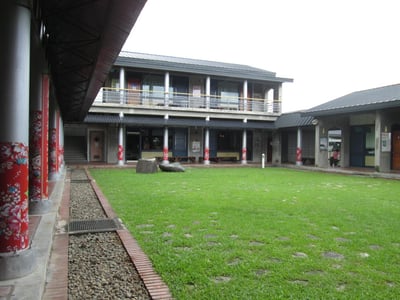 붉은 꽃무늬 기둥이 인상적인 회랑으로 둘러싸인 넓은 잔디 마당과 현대적인 2층 석조 건물의 평화로운 풍경입니다.