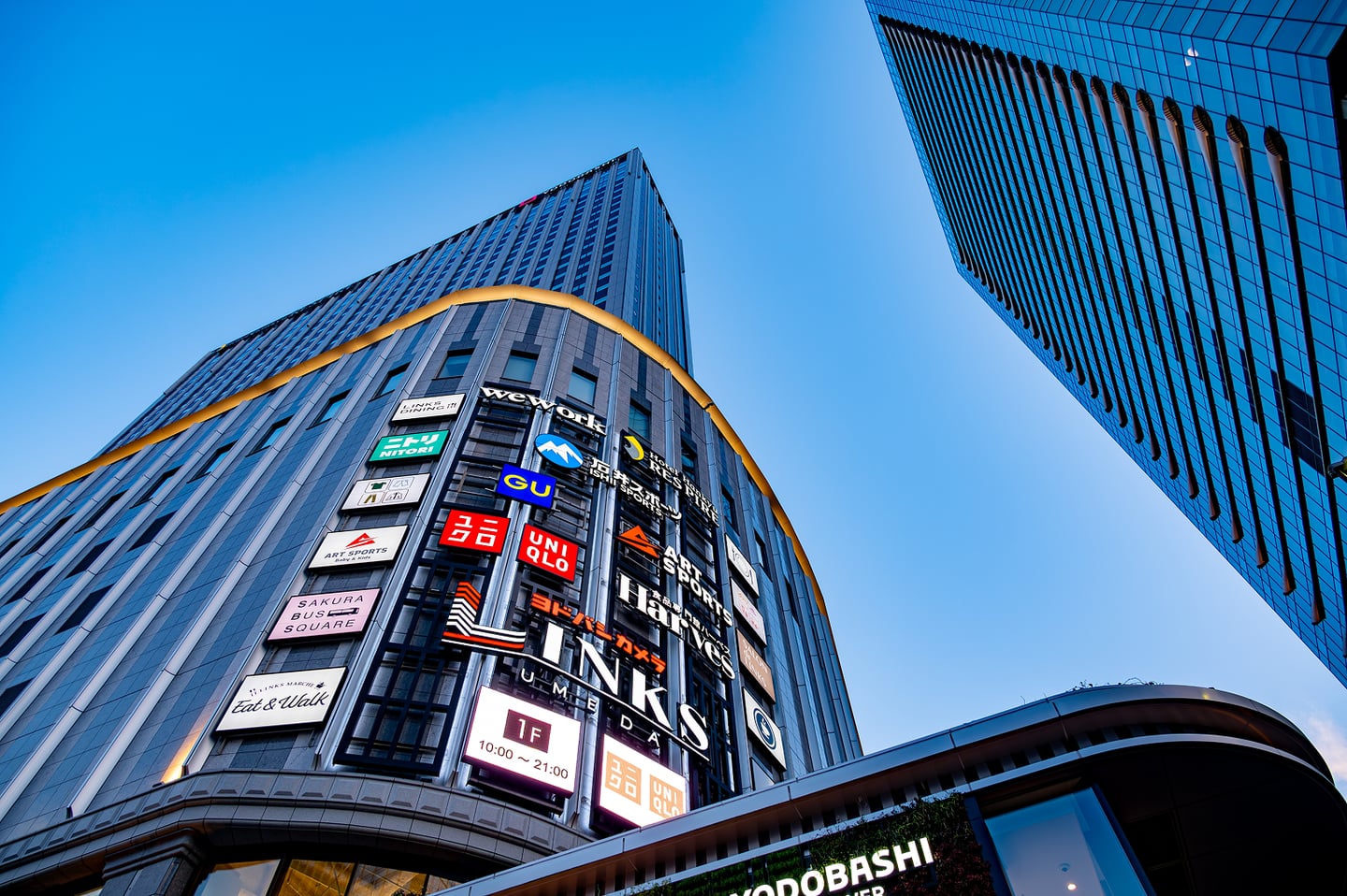 푸른 하늘 아래 유니클로와 요도바시 카메라 등 여러 브랜드 간판이 화려하게 걸려 있는 오사카 링크스 우메다 빌딩의 웅장한 외관입니다.