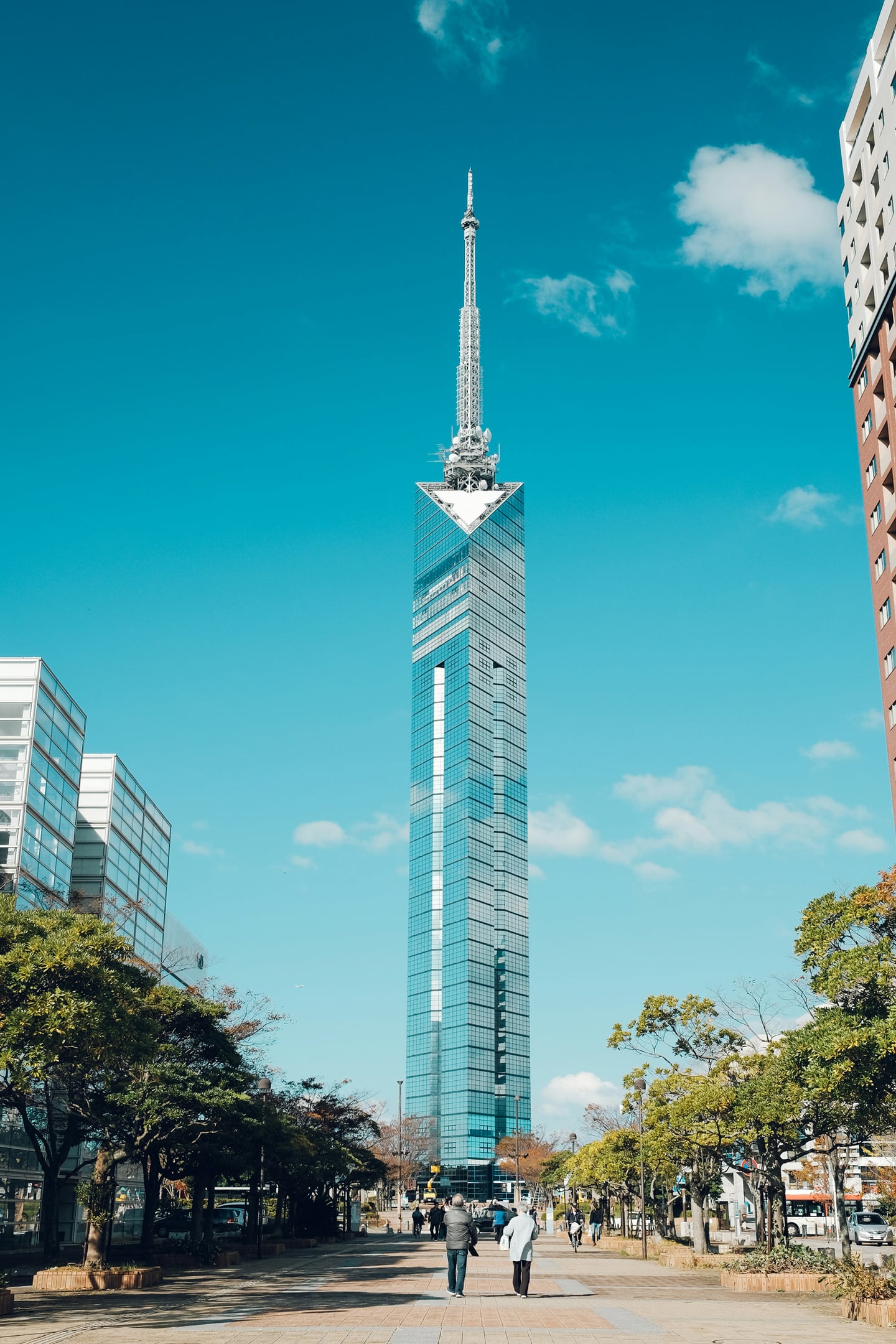 쾌청한 날씨 속에서 푸른 유리 외벽이 빛나는 후쿠오카 타워와 그 아래로 이어지는 평화로운 도심 산책로의 풍경입니다.