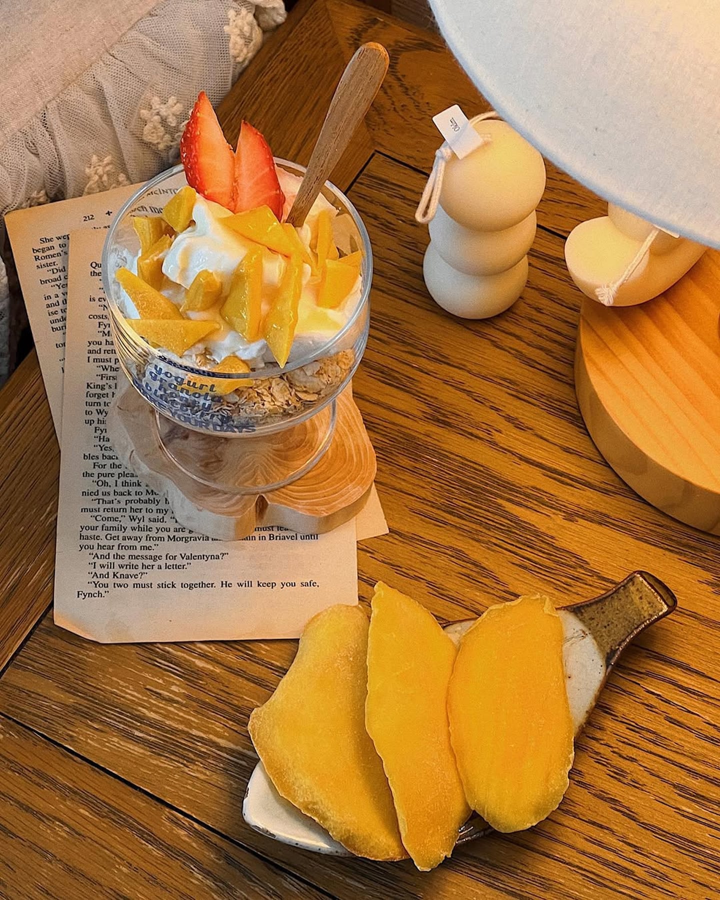 따뜻한 조명 아래 나무 테이블 위로 놓인 신선한 과일 요거트 볼과 말린 망고 슬라이스가 담긴 접시가 감성적인 홈카페 분위기를 연출합니다.