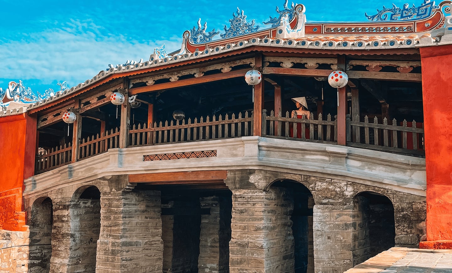 푸른 하늘 아래 베트남 호이안 내원교의 고풍스러운 목조 구조와 붉은 외벽, 그리고 전통 모자를 쓴 인물이 어우러진 풍경입니다.