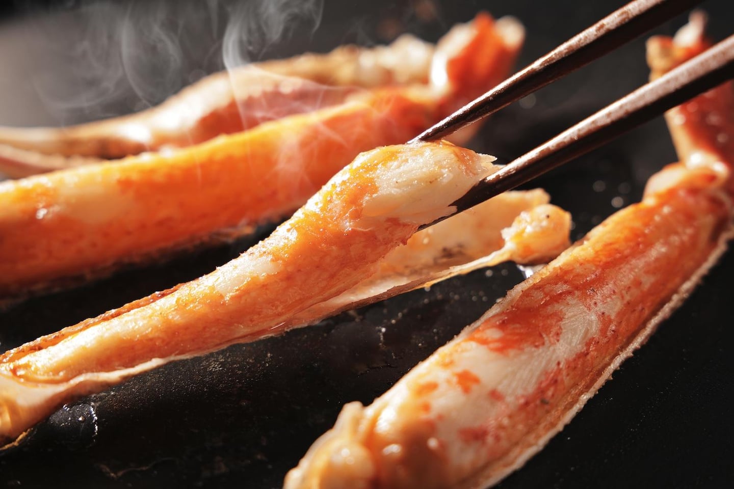 뜨거운 김이 올라오는 불판 위에서 젓가락으로 집어 올린 통통하고 붉은 대게 다리 살의 클로즈업 이미지입니다.