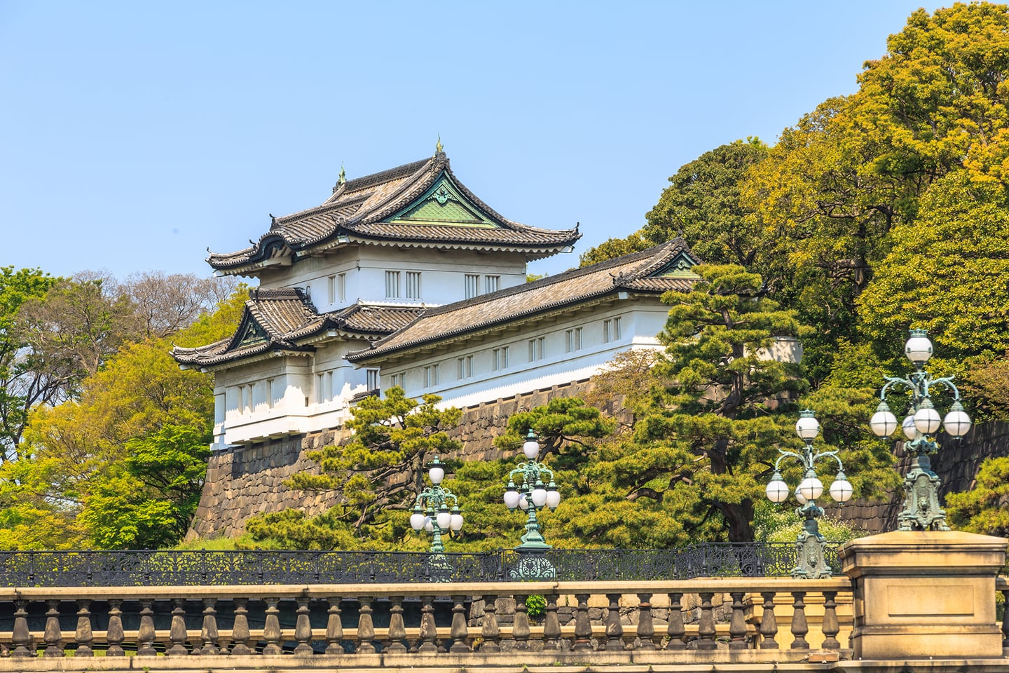 도쿄 황궁의 전통적인 백색 망루가 거대한 돌담 위에 세워져 있고, 그 앞으로 화려한 장식의 가로등과 푸른 정원이 조화를 이루는 풍경입니다.