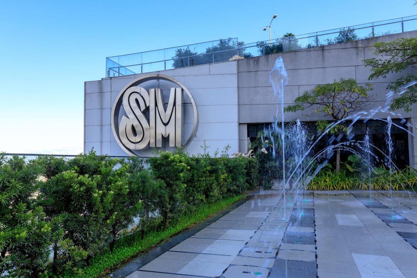 맑은 날씨 속 SM 쇼핑몰 건물의 은색 로고와 바닥에서 솟아오르는 분수대가 있는 야외 테라스 광장의 풍경입니다.