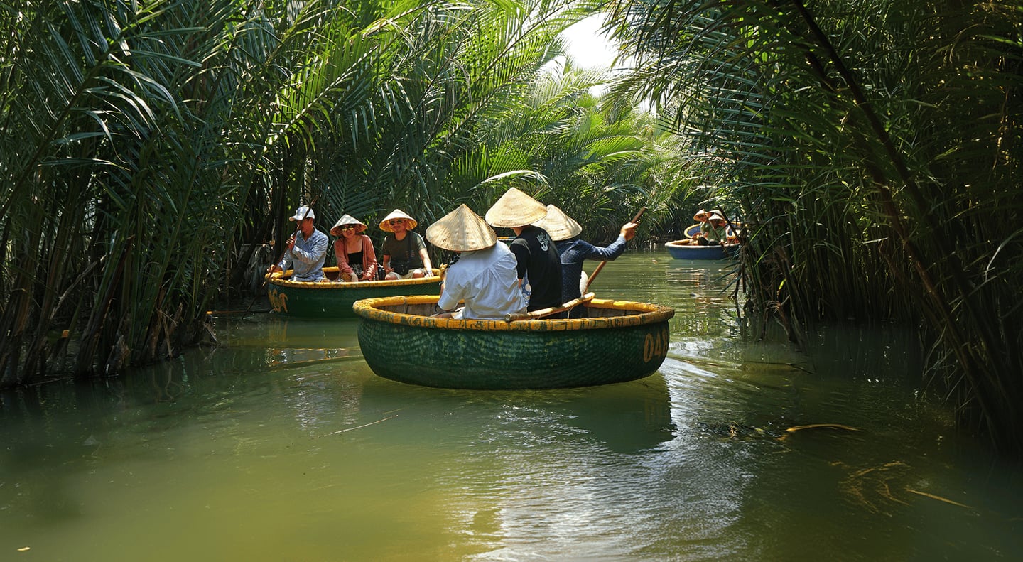 울창한 야자수 숲이 우거진 베트남 호이안의 좁은 수로에서 전통 모자인 농라를 쓴 관광객들이 둥근 바구니 배를 타고 평화롭게 노를 저으며 이동하는 풍경입니다.