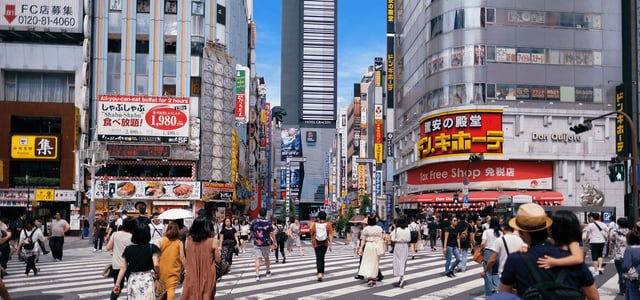 도쿄 신주쿠 가부키초 거리에서 횡단보도를 건너는 많은 사람들과 돈키호테 매장, 멀리 보이는 고질라 조형물이 특징인 도심 풍경입니다.