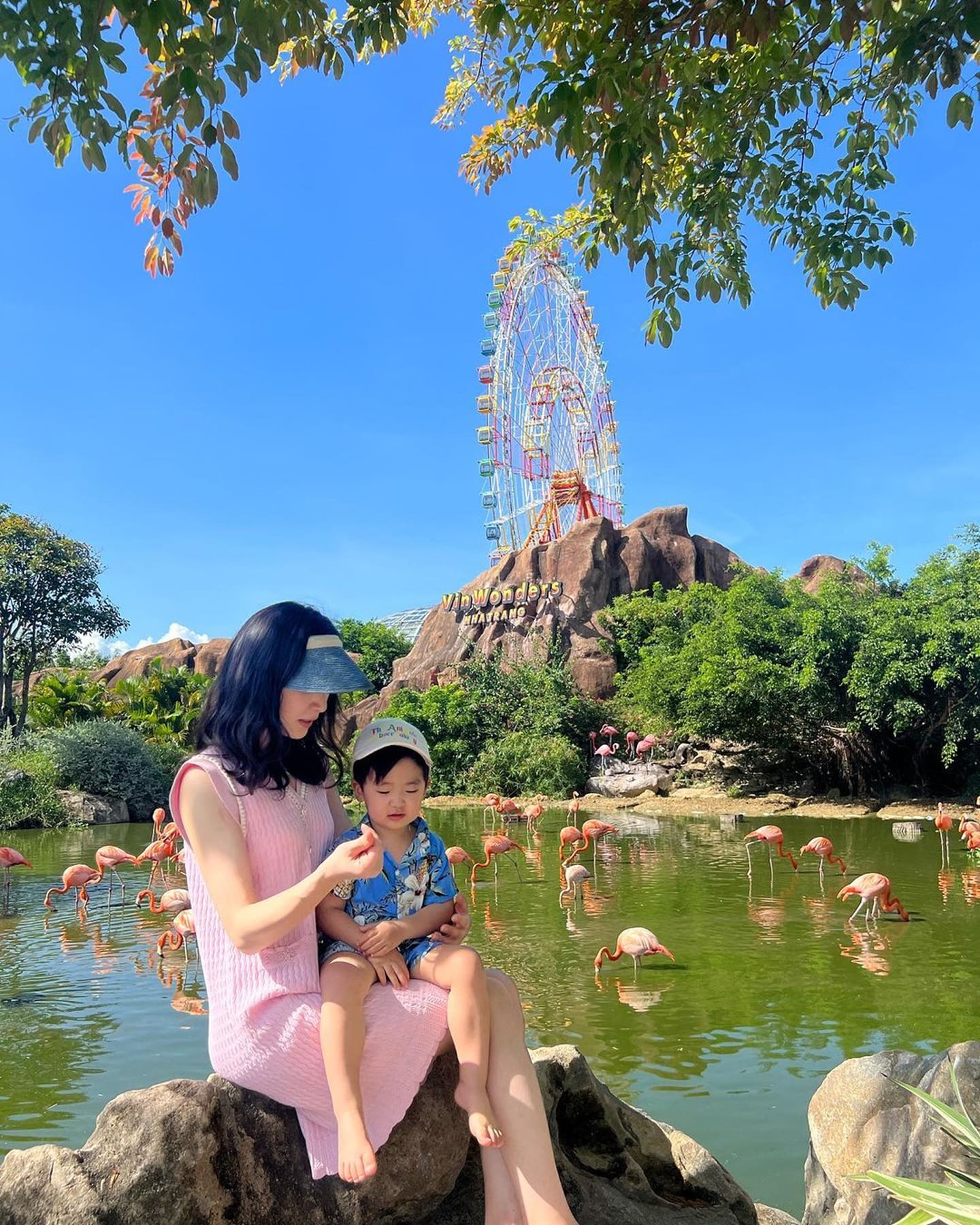 베트남 나트랑 빈원더스 테마파크에서 대관람차와 홍학 무리가 보이는 호수를 배경으로 다정하게 앉아 있는 엄마와 어린 아들의 모습입니다.