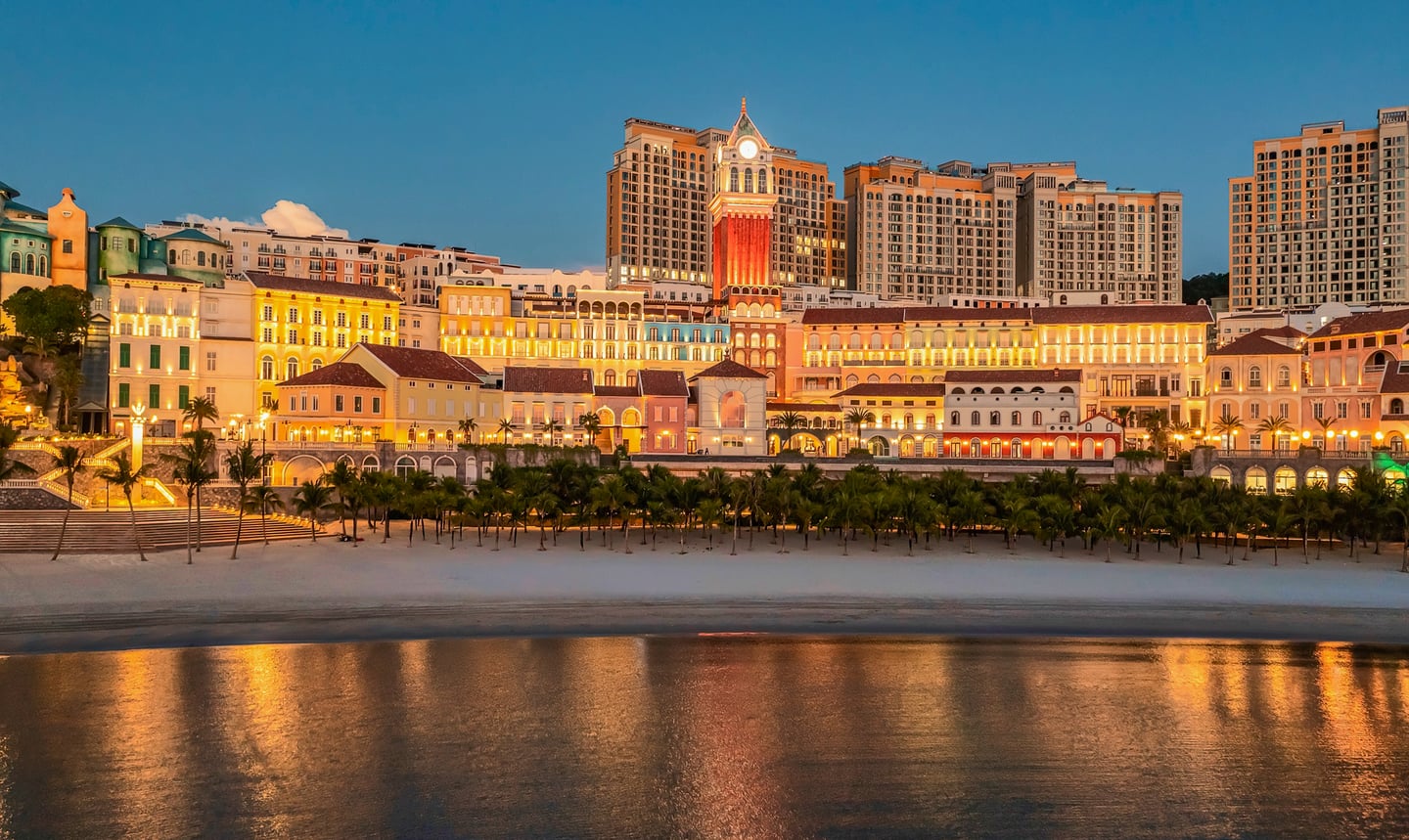 해변을 따라 늘어선 화려한 조명의 유럽풍 건물들과 시계탑이 바다에 반사되는 푸꾸옥 선셋 타운의 낭만적인 밤 풍경입니다.