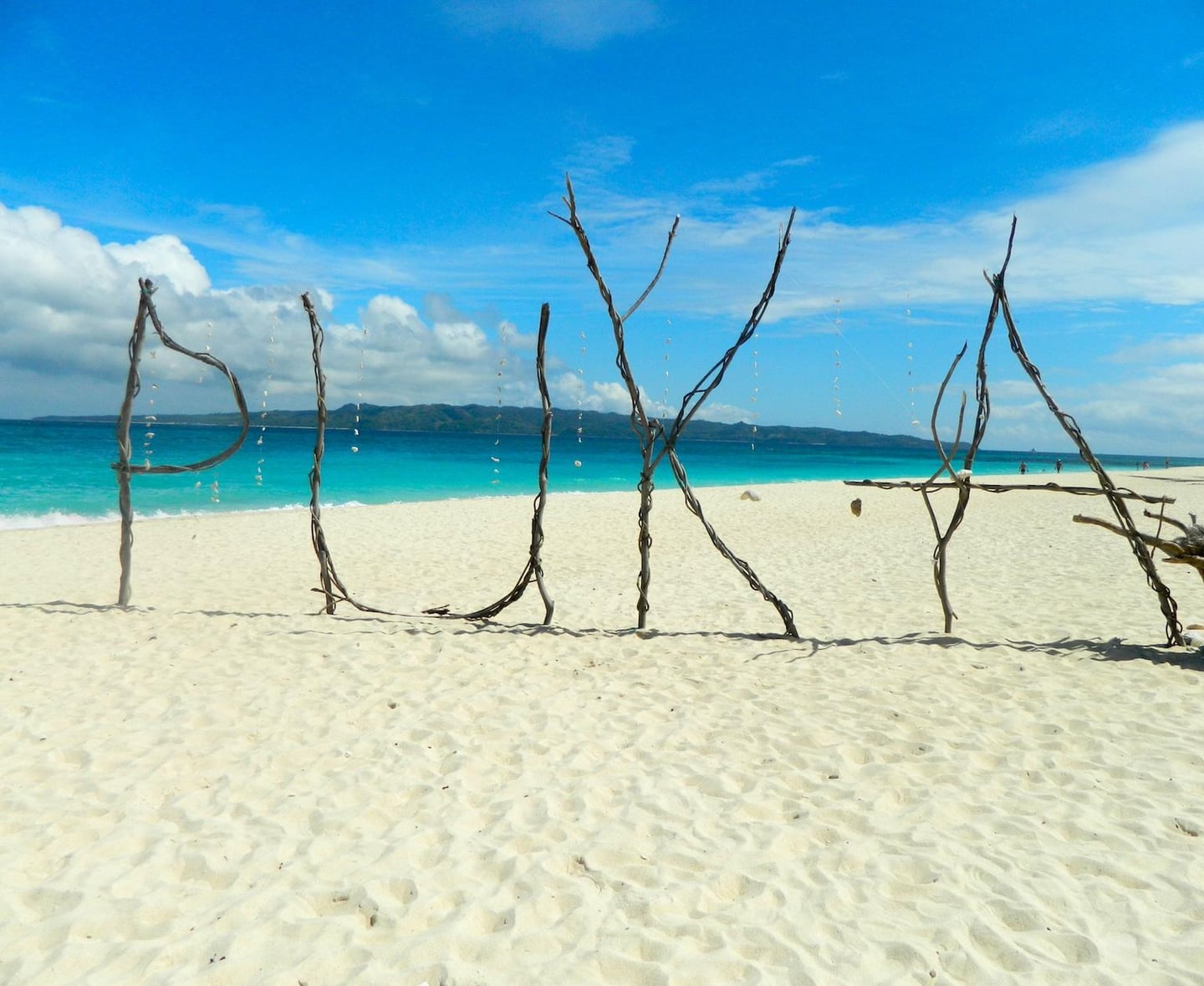 보라카이 푸카 쉘 비치의 하얀 모래사장 위에 나뭇가지로 만든 커다란 푸카 글자 조형물이 에메랄드빛 바다를 배경으로 서 있는 풍경입니다.