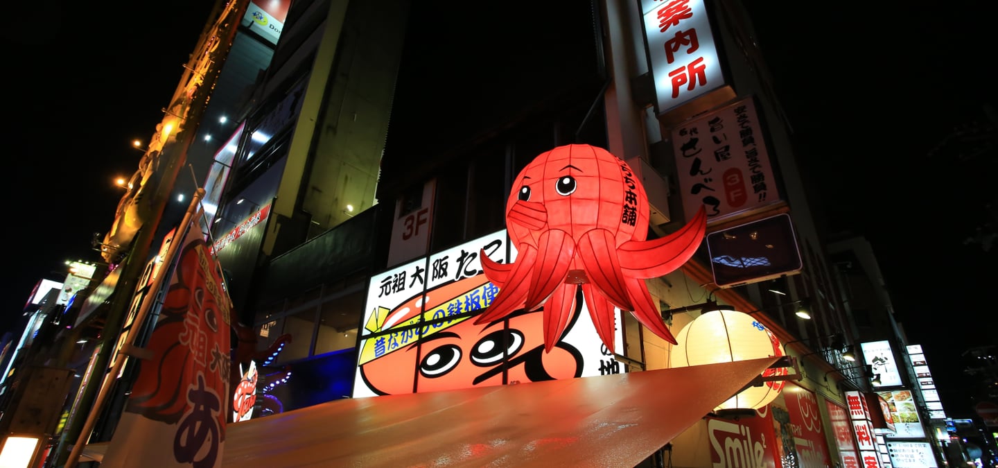 오사카 도톤보리의 밤거리에서 화려한 조명을 받으며 시선을 사로잡는 거대한 붉은 문어 입체 간판과 일본어 네온사인들이 가득한 풍경입니다.