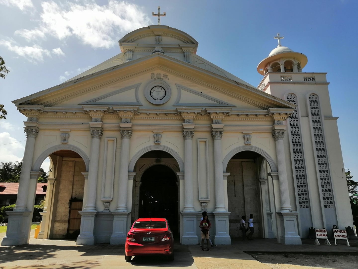 웅장한 기둥과 시계탑이 돋보이는 흰색 석조 성당 건물 앞에 빨간색 자동차가 세워져 있는 맑은 날의 전경입니다.