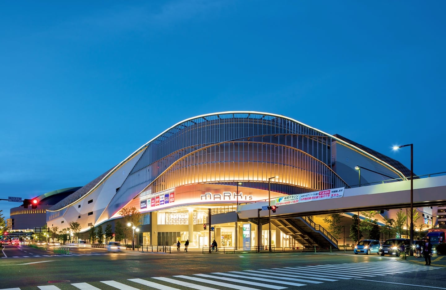 어스름한 저녁 시간대, 부드러운 곡선형 지붕과 따뜻한 오렌지빛 조명이 어우러진 현대적인 대형 쇼핑몰 건물이 도심 교차로 앞에 자리하고 있습니다.