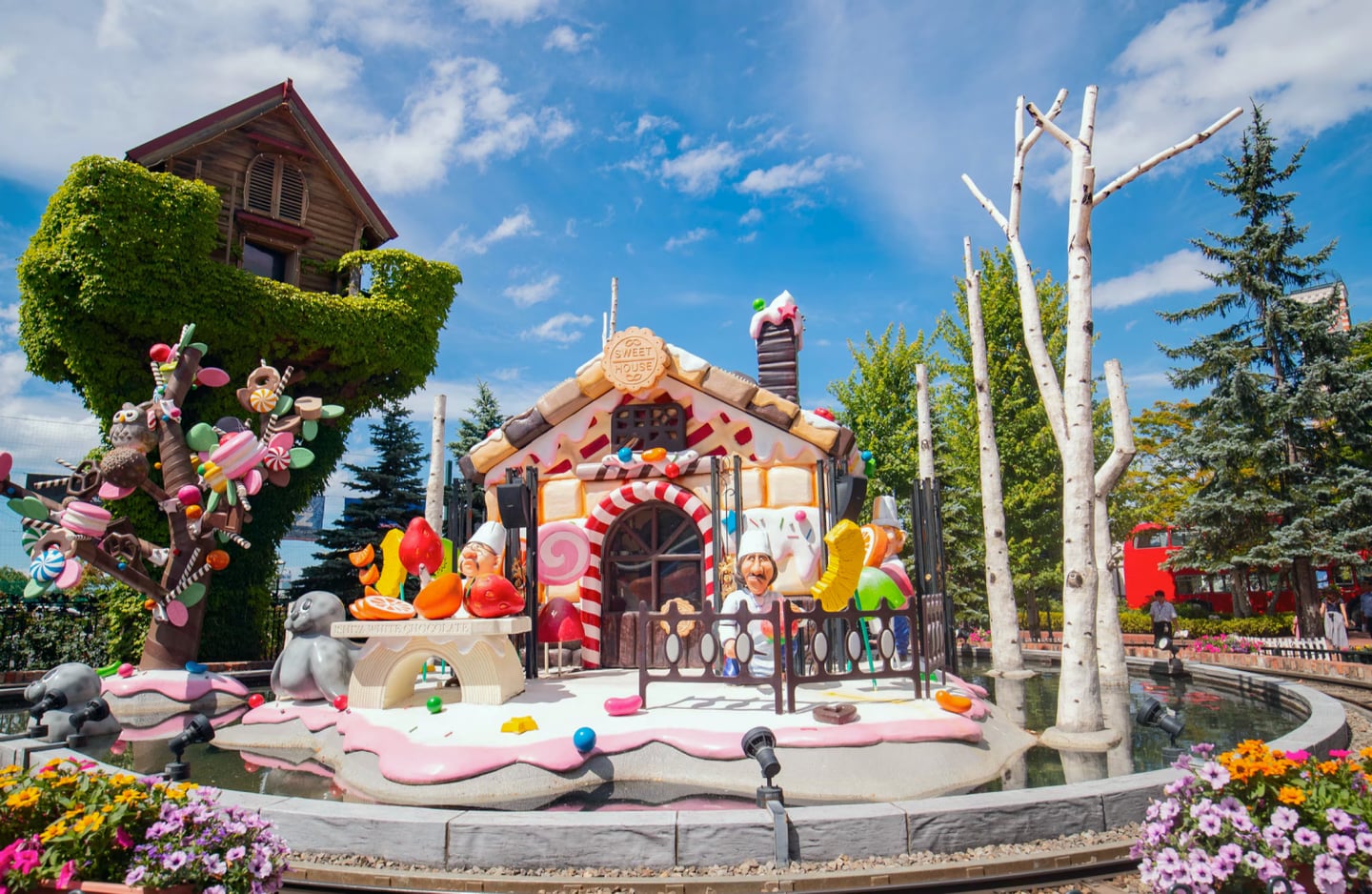맑은 날 야외 테마파크 연못 위에 세워진 화려한 과자 집 조형물과 사탕 나무가 어우러진 동화 같은 풍경입니다.