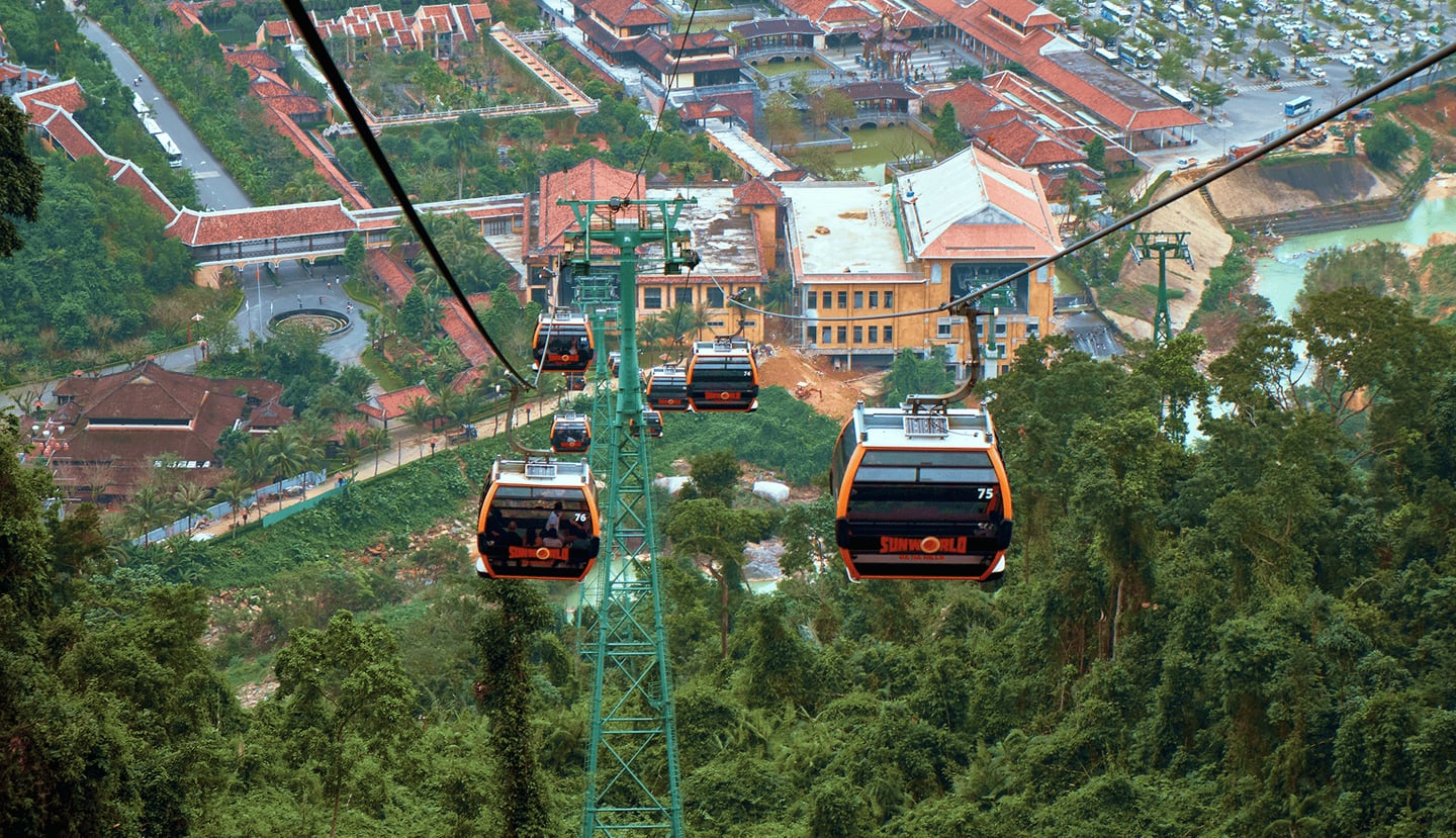 푸른 숲이 우거진 산등성이 위로 오렌지색 선월드 케이블카들이 줄지어 이동하며 멀리 붉은 기와지붕의 마을과 광장이 내려다보이는 풍경입니다.