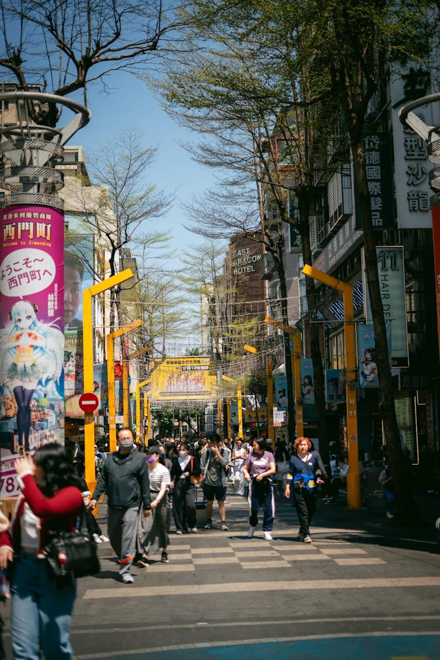 타이베이 시먼딩의 활기찬 보행자 거리에서 다양한 상점 간판과 노란색 구조물 사이로 많은 사람들이 산책을 즐기는 풍경입니다.