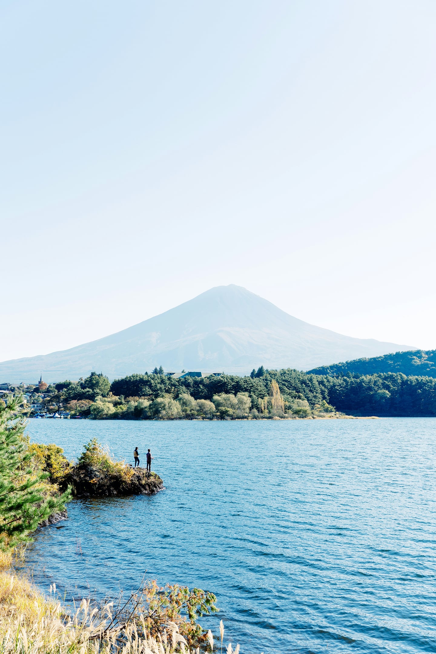 맑고 푸른 호수와 울창한 숲 너머로 거대한 후지산이 배경으로 펼쳐진 가운데 낚시를 즐기는 듯한 두 사람의 실루엣이 담긴 평온한 자연 풍경입니다.