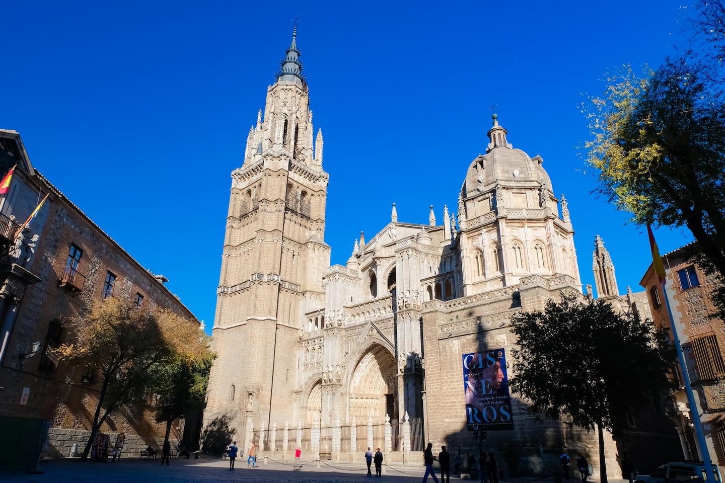 스페인 톨레도 대성당의 정교한 석조 외관과 높은 첨탑이 맑은 하늘과 어우러진 풍경으로, 광장을 지나는 사람들의 모습이 평화로운 분위기를 자아냅니다.