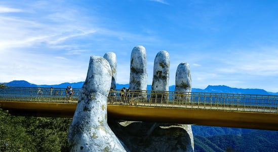 푸른 하늘과 산맥을 배경으로 거대한 두 손이 황금빛 보행교를 떠받치고 있는 베트남 다낭 바나힐 골든 브릿지의 웅장한 전경입니다.