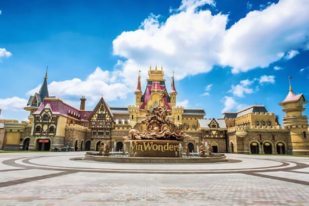 맑고 푸른 하늘 아래 빈원더스 로고가 새겨진 화려한 조각 분수와 동화 속 성처럼 꾸며진 테마파크 건물이 어우러진 이국적인 광장 풍경입니다.
