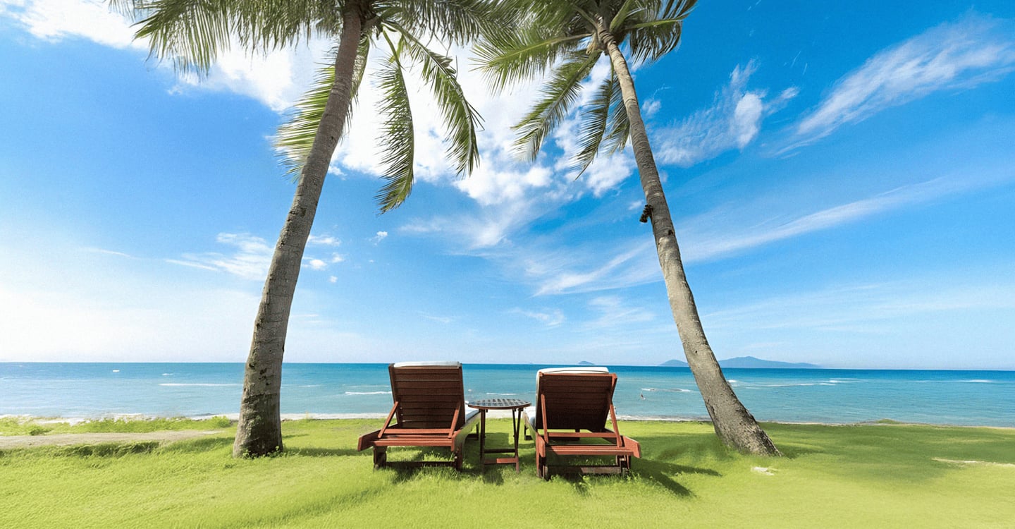 에메랄드빛 바다가 펼쳐진 해변가 잔디밭 위로 야자수 두 그루와 나란히 놓인 나무 선베드가 평화로운 휴양지의 정취를 자아냅니다.