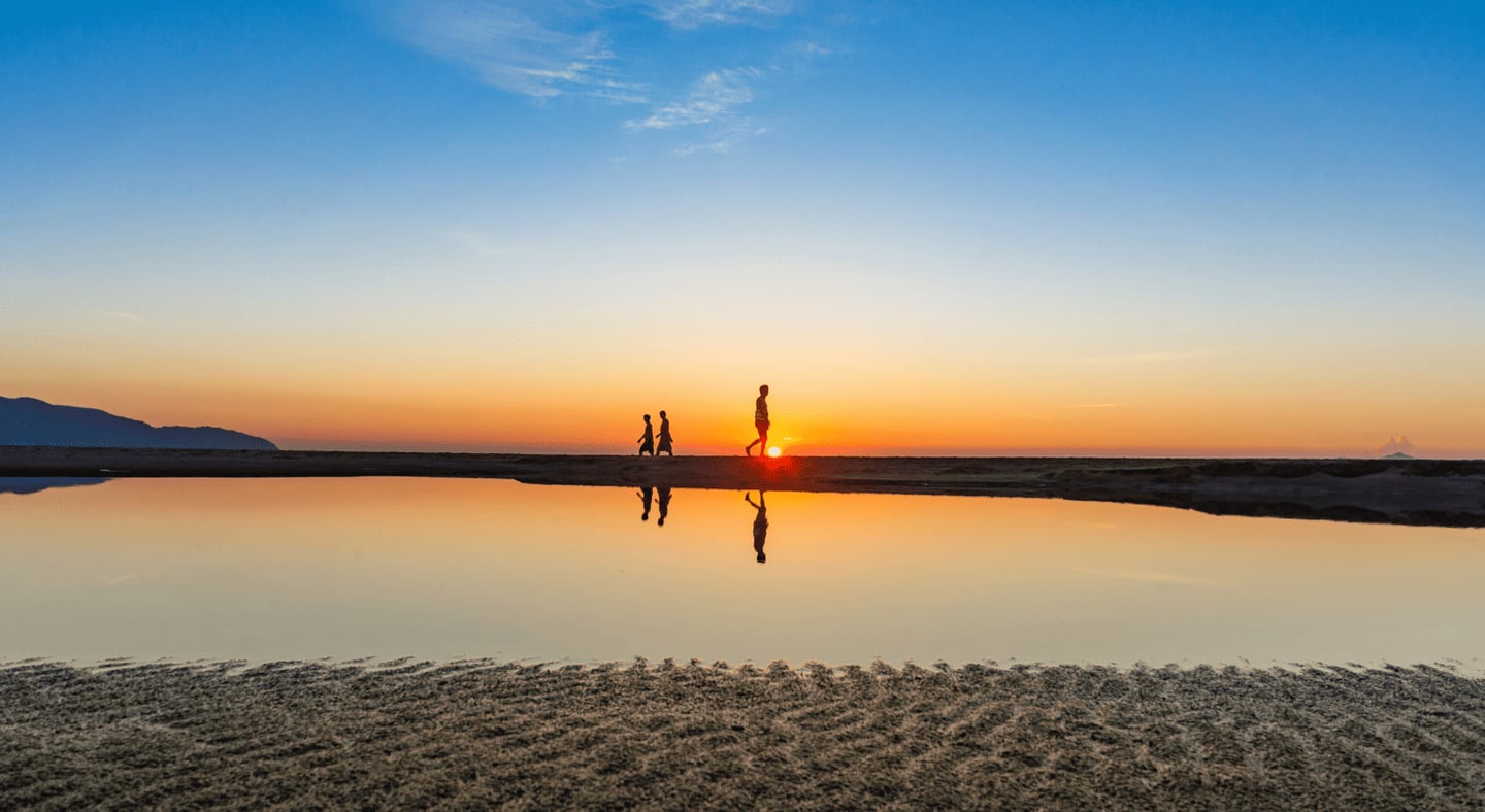 노을이 지는 해변의 수평선을 따라 걷는 사람들의 실루엣과 물결 없는 수면에 비친 아름다운 반영이 돋보이는 평온한 저녁 풍경입니다.