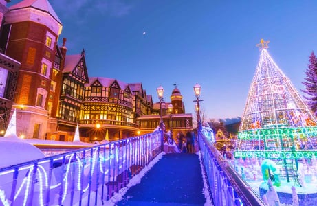 눈 내린 겨울 저녁 화려한 전구 장식으로 빛나는 대형 트리와 고풍스러운 붉은 벽돌 건물이 어우러진 환상적인 야경 모습입니다.