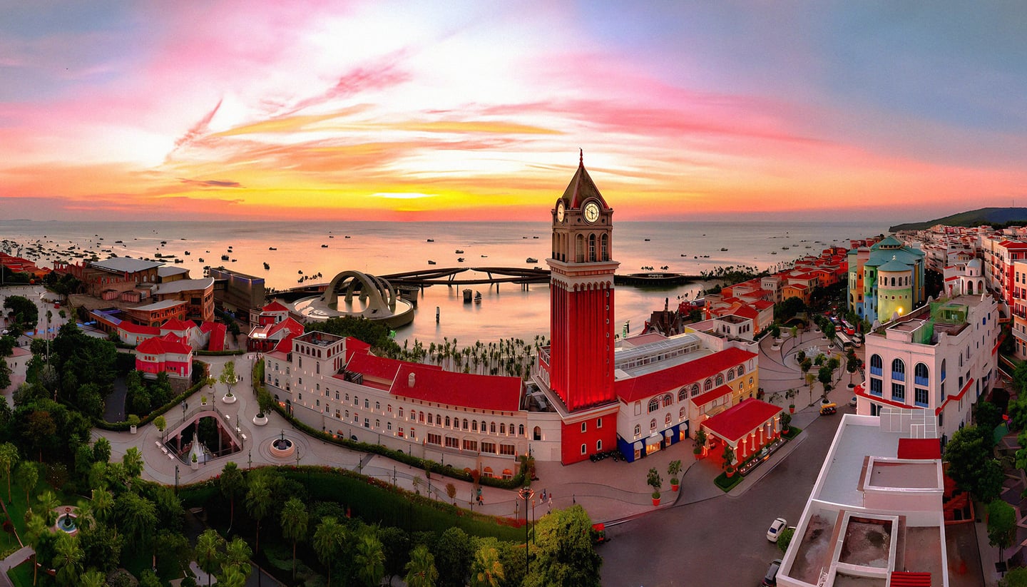 노을이 지는 바닷가 마을에 우뚝 솟은 붉은 시계탑과 유럽풍 건물들이 어우러져 환상적인 저녁 야경을 선사하는 풍경입니다.