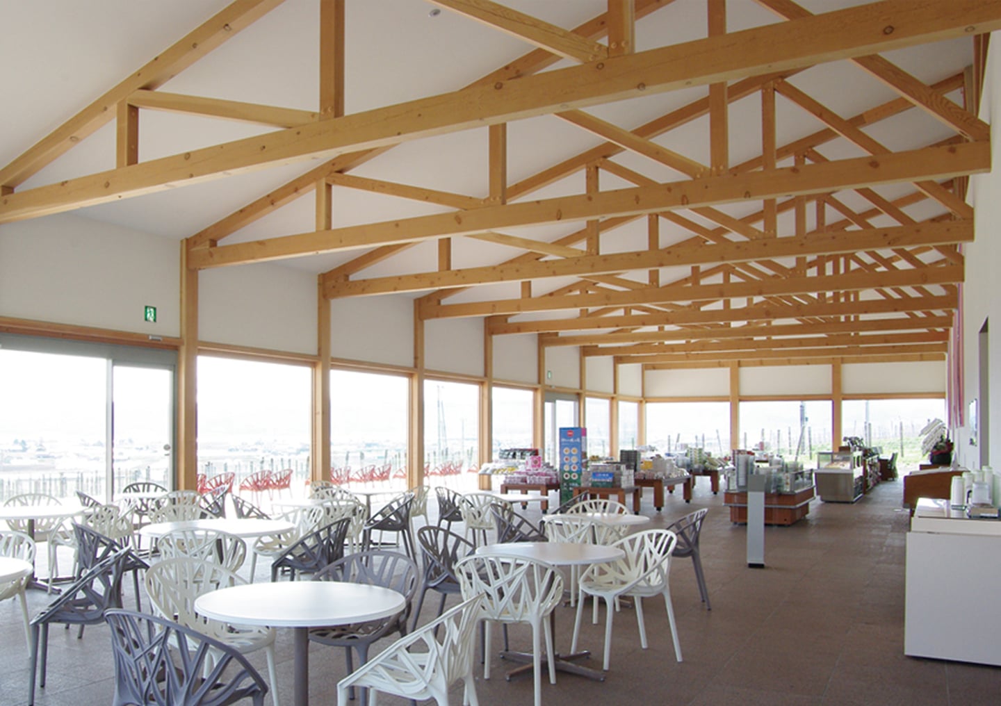 나무 서까래가 드러난 높은 천장 아래 세련된 디자인의 의자와 테이블이 놓인 넓은 카페 내부 전경으로 창밖의 풍경이 시원하게 펼쳐집니다.