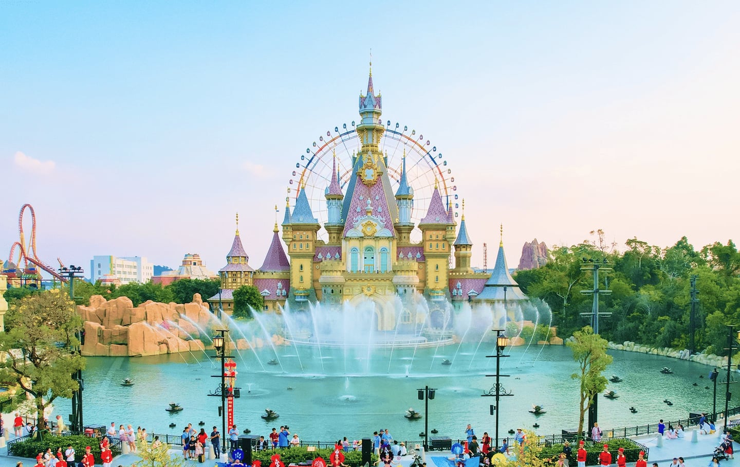 동화 속 성과 대관람차가 보이는 테마파크 호수에서 시원하게 뿜어져 나오는 분수 쇼와 나들이를 즐기는 사람들의 활기찬 모습입니다.