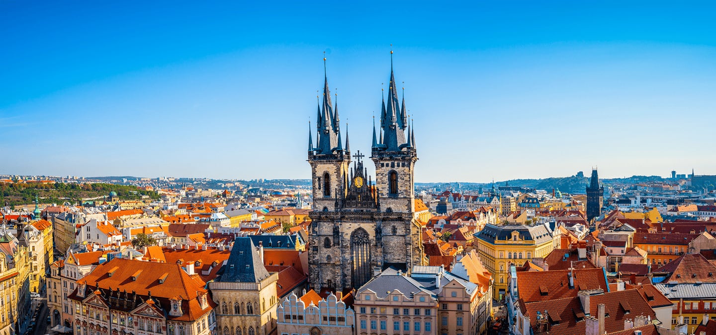 푸른 하늘 아래 우뚝 솟은 프라하 틴 성모 교회의 검은 첨탑과 구시가지의 붉은 지붕들이 조화를 이루는 이국적인 도시 경관입니다.