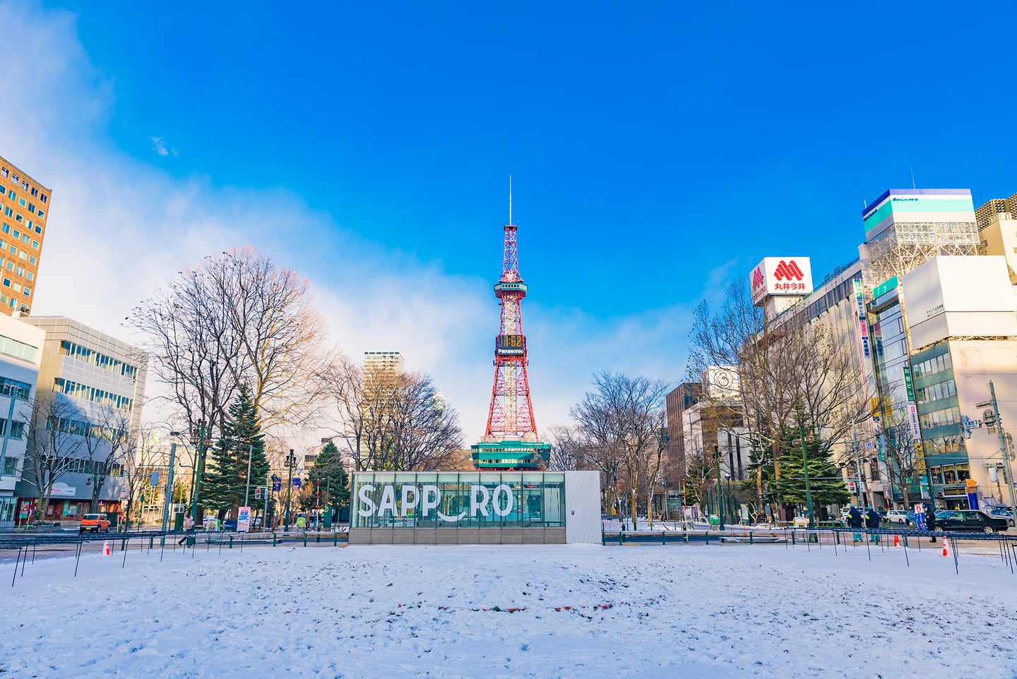흰 눈이 쌓인 광장 뒤로 붉은색 삿포로 TV 타워가 선명하게 보이고, 현대적인 빌딩들과 앙상한 겨울 나무들이 어우러진 맑은 날의 삿포로 시내 풍경입니다.