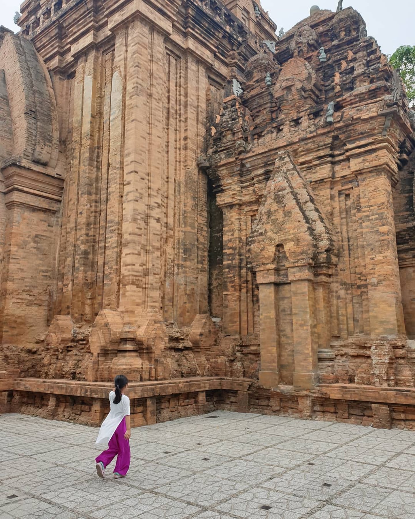 나트랑 포나가르 첨탑의 거대한 붉은 벽돌 건축물 아래에서 흰색 상의와 보라색 바지를 입은 여행객이 광장을 가로질러 걸어가고 있습니다.