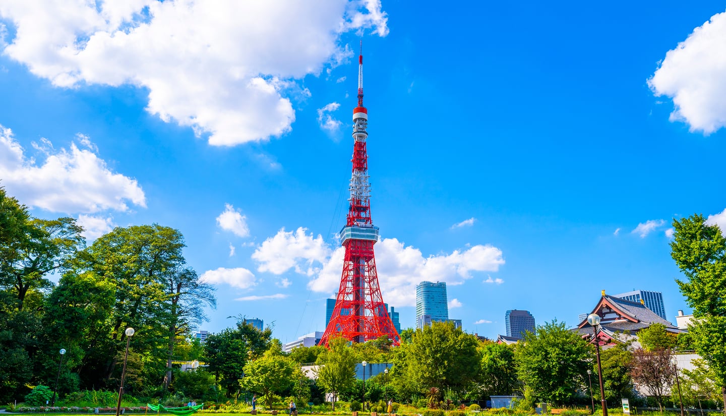 맑고 푸른 하늘 아래 선명한 오렌지빛 도쿄 타워가 푸른 나무들 사이로 솟아 있으며 주변에는 현대적인 고층 빌딩과 전통 가옥이 조화를 이룹니다.