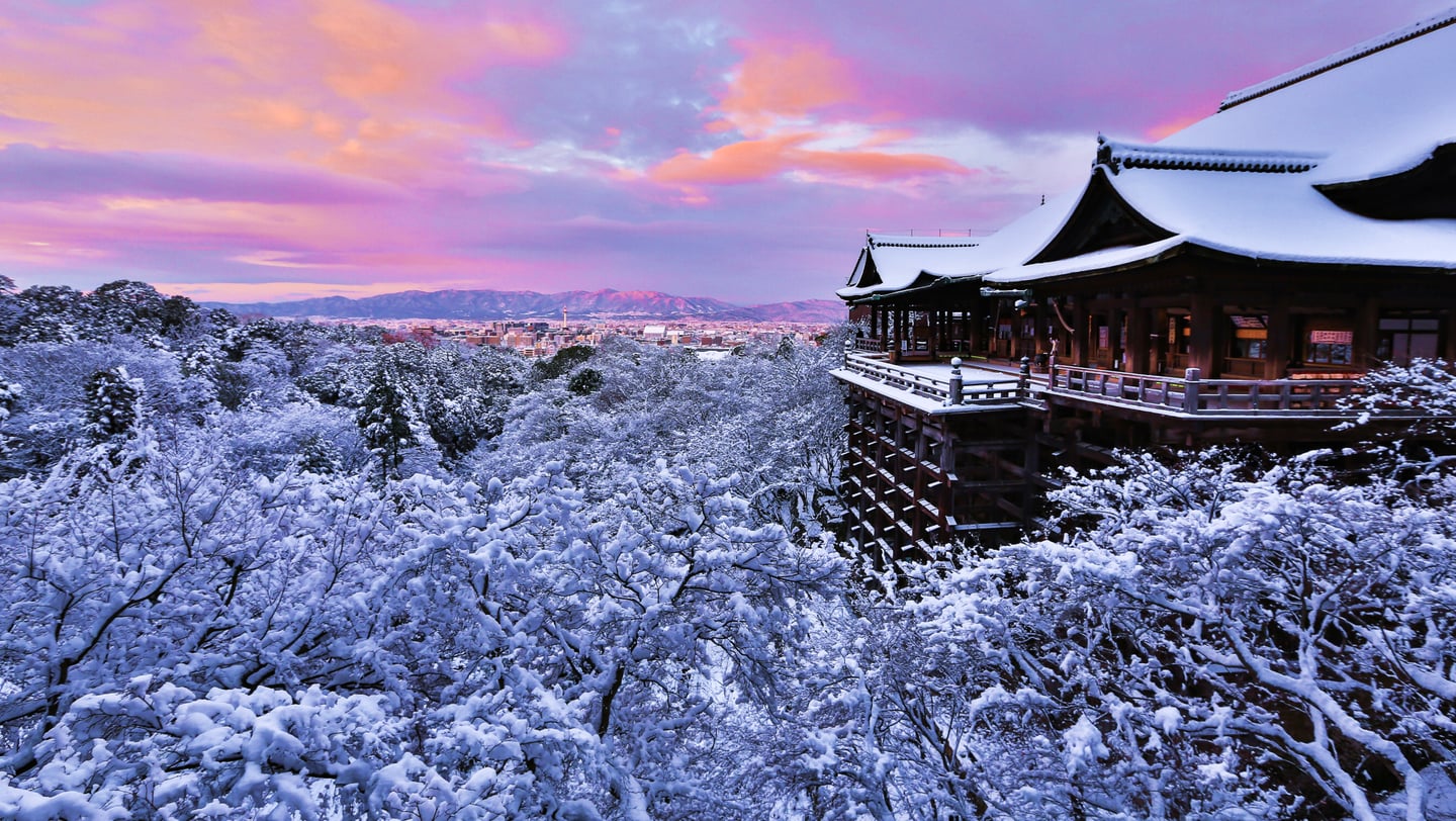 보랏빛 노을이 지는 하늘 아래 하얀 눈이 소복이 쌓인 일본 교토 기요미즈데라의 웅장한 목조 건축물과 겨울 숲의 전경입니다.