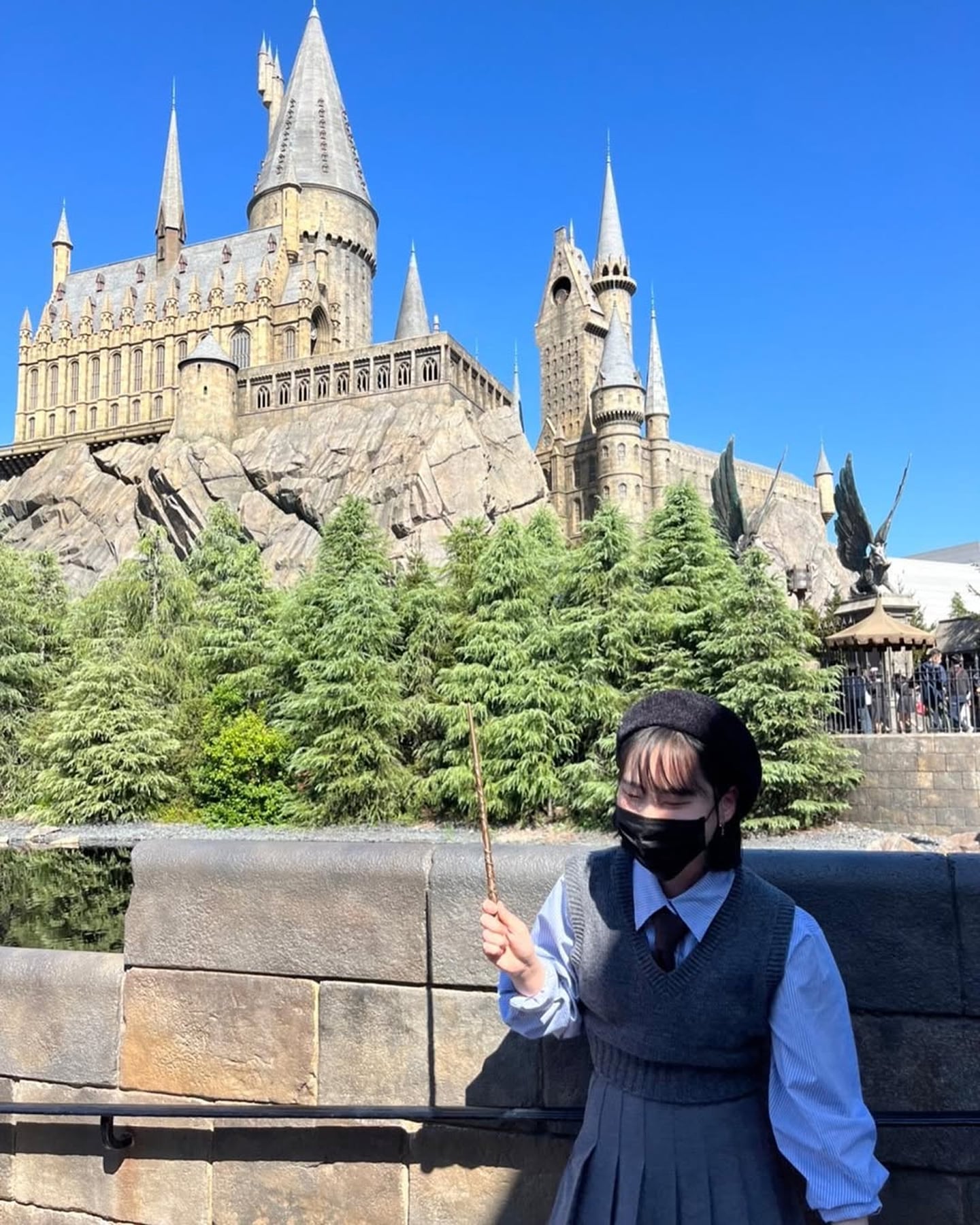푸른 하늘 아래 웅장하게 솟은 호그와트 성을 배경으로 마법 지팡이를 든 여성이 교복 차림으로 서 있는 유니버설 스튜디오의 풍경입니다.