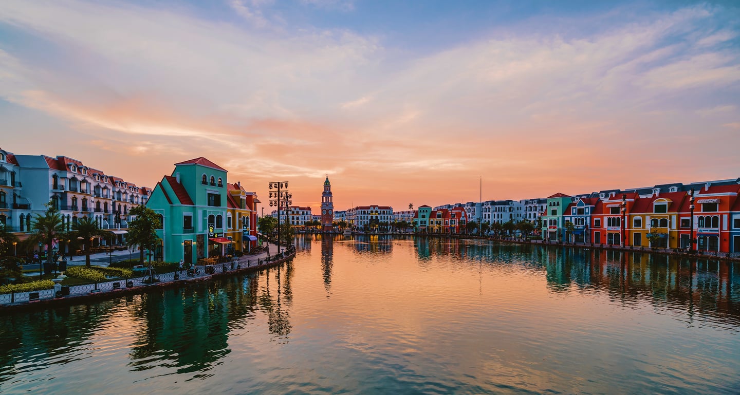 노을빛으로 물든 하늘 아래 운하를 따라 늘어선 파스텔 톤의 유럽풍 건축물들과 물 위에 비친 평화로운 도시의 반영 모습입니다.